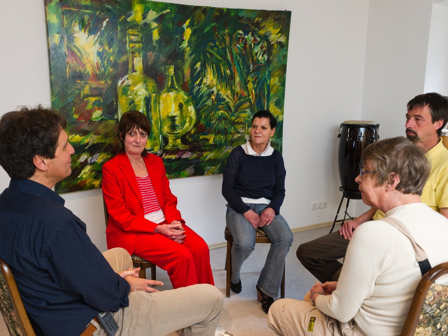 Fünf Menschen sitzen im Kreis auf Stühlen einander zugewandt. Die Atmosphäre ist konzentriert und ernst. Im Hintergrund hängt ein Gemälde eines Dschungels mit zwei Glasflaschen.