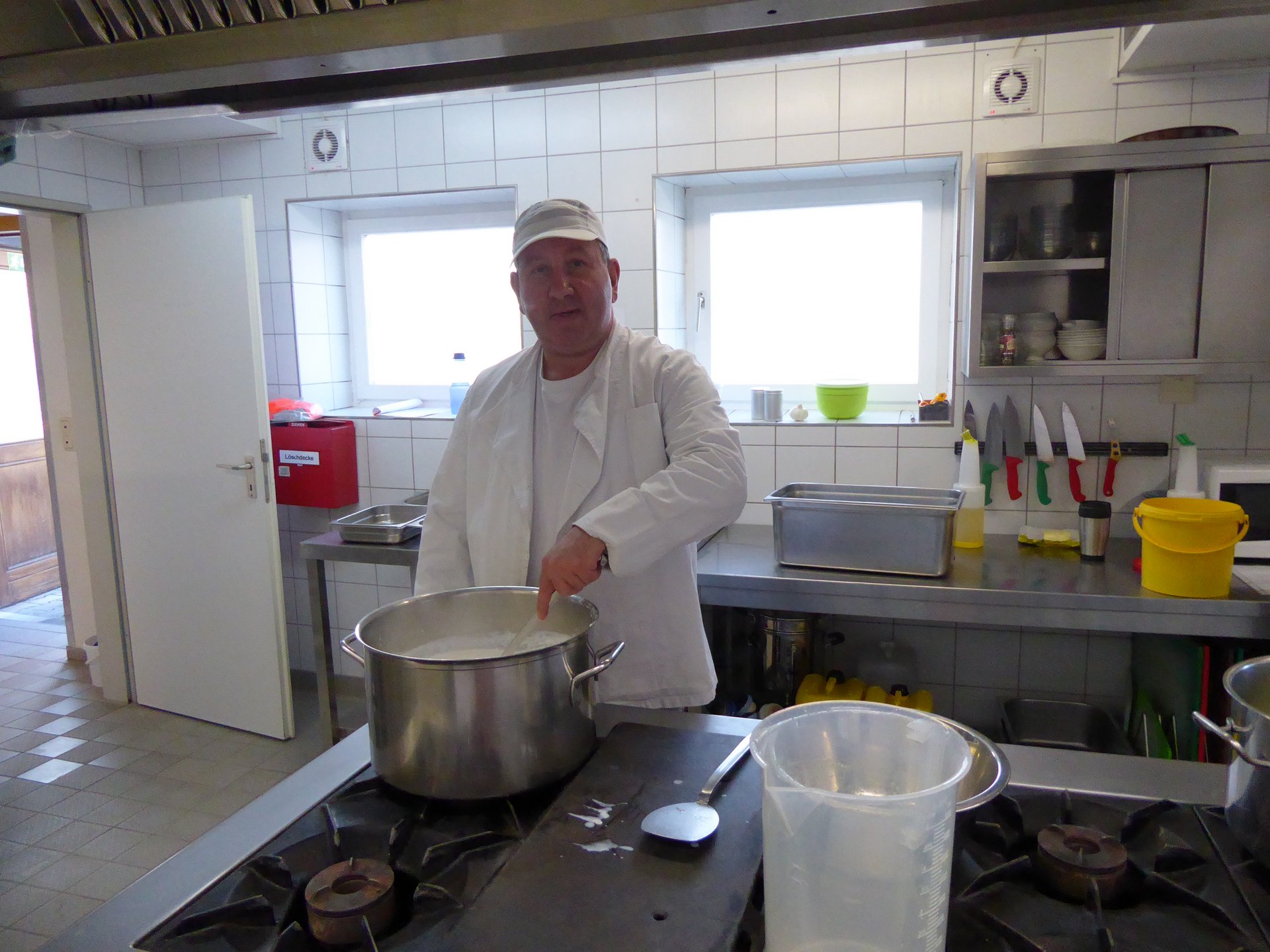 In einer Küche steht ein weiß gekleideter Mann mit Kochmütze und rührt in einem Topf.
