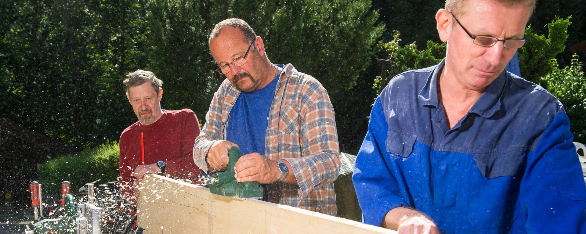 Zwei Männer halten ein Brett fest, während ein dritter mit der Hobelmaschine die Kante bearbeitet. Späne fliegen, alle drei wirken konzentriert.