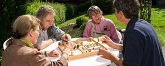 Vier Menschen sitzen draußen um einen kleinen runden Tisch und spielen Schach. Sie unterhalten sich angeregt.