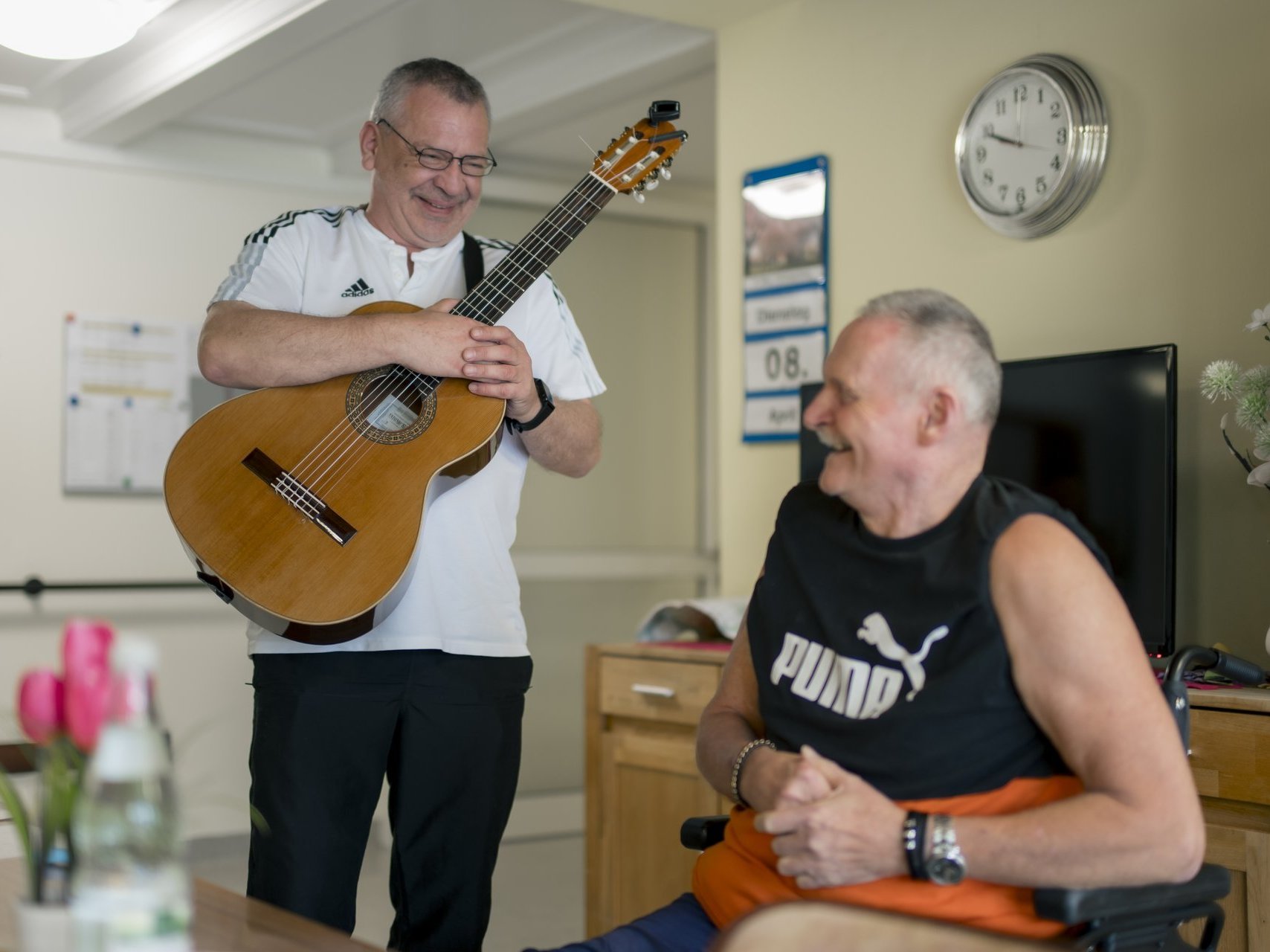 Ein Herr steht mit einer Gitarre neben einem anderen, der im Rollstuhl sitzt und beide lachen.