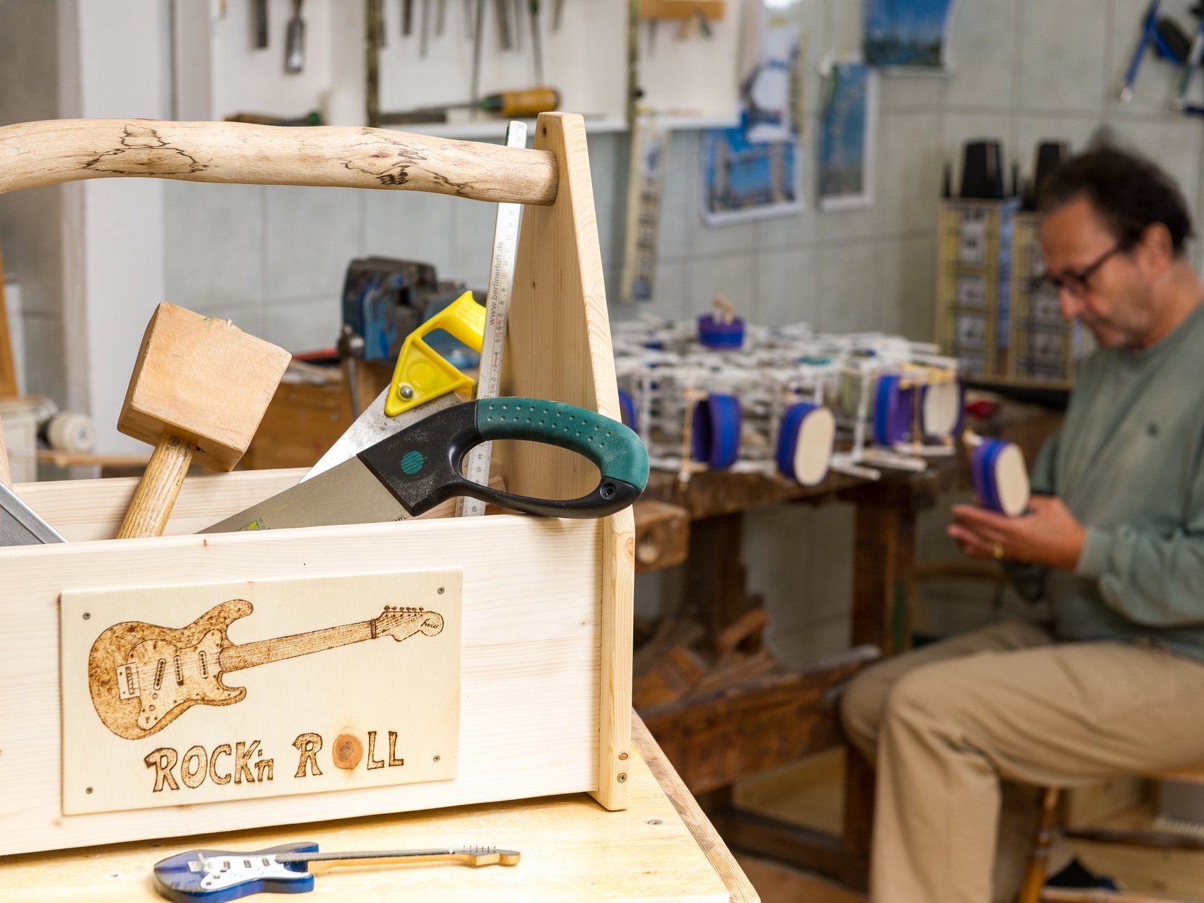 Eine Holzwerkstatt. Im Vordergrund steht eine Werkzeugkiste in der verschiedene Werkzeuge zu sehen sind. Im Hintergrund arbeitet ein Mann an einem Werkstück.