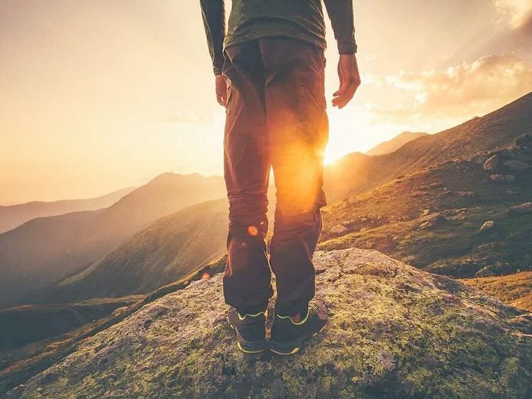 Eine Person, zu sehen von Hinten, steht auf einem Felsen. Im Hintergrund die untergehende Sonne