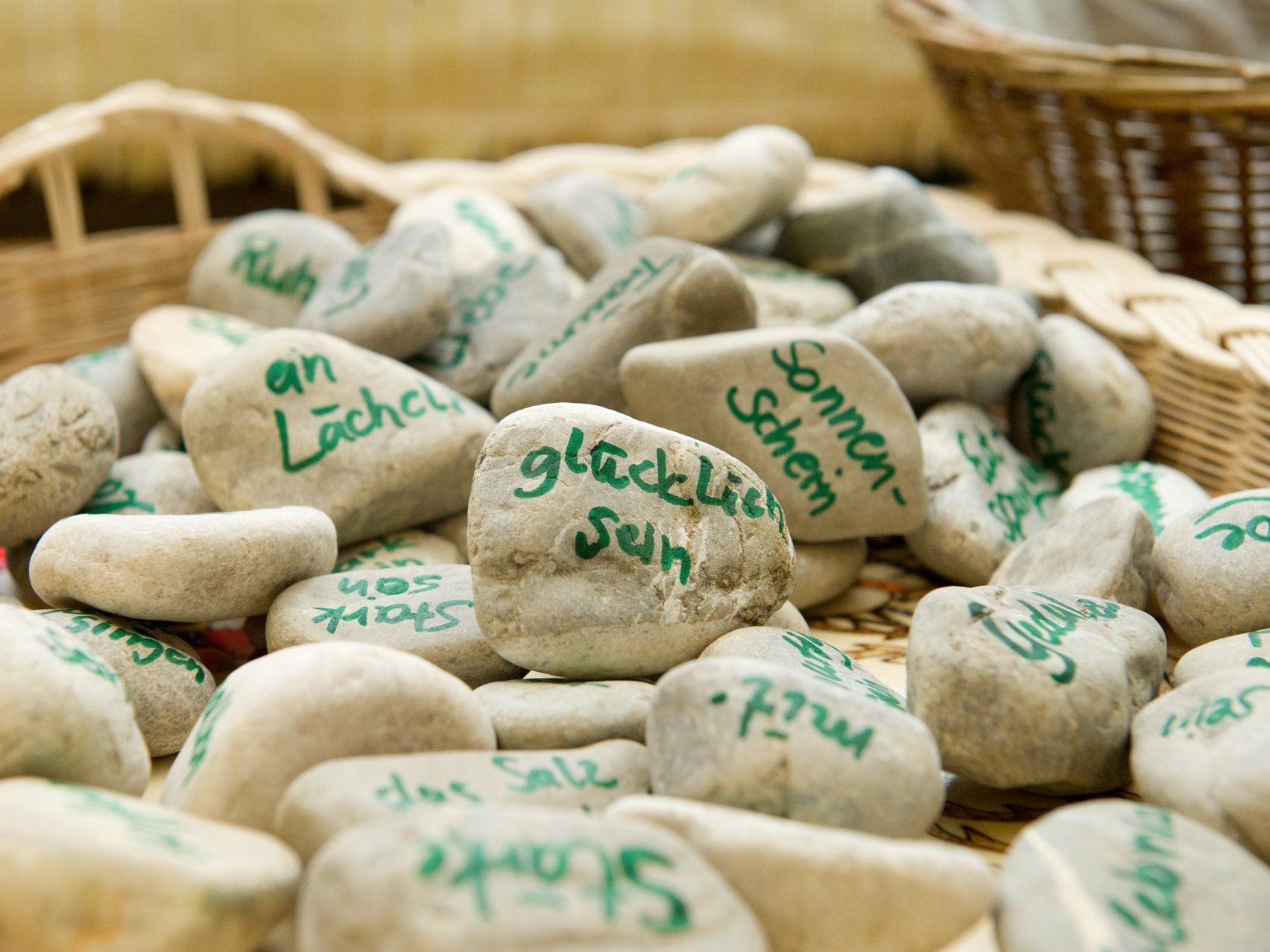 In einem Korb liegen viele hellgraue Flusskiesel auf denen mit grüner Farbe Worte geschrieben stehen wie lächeln, glücklich sein, sonnenschein