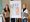Zwei Frauen stehen lächelnd auf beiden Seiten eines Flipchart-Ständers in einem Raum mit Holzboden. Auf dem Flipchart ist ein großes Blatt mit Text und farbigen Abschnitten (grün und orange) befestigt. Im Hintergrund ist eine Tafel zu erkennen.