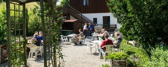 In einer Aussenanlage vor einem Haus stehen einige Tische mit Stühlen auf denen mehrere Menschen sitzen, sich unterhalten oder lesen. Die Sonne scheint.